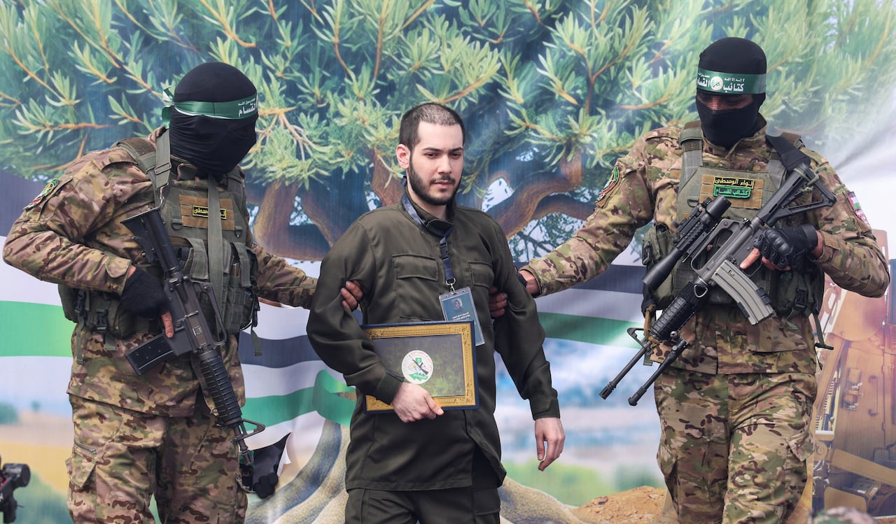 Two people wearing military uniforms, holding rifles with their faces covered, hold the arms of a civilian man.