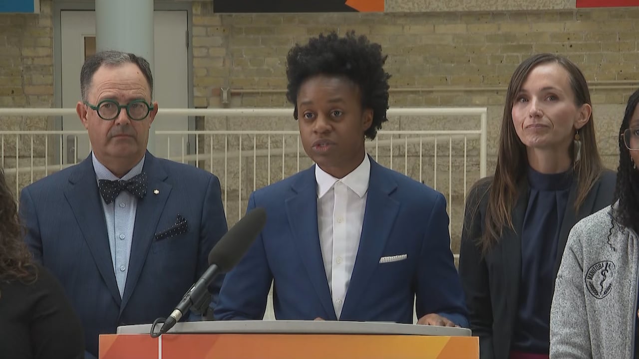A person with dark hair wears a blue suit and speaks into a microphone at an orange podium. They are surrounded by other people