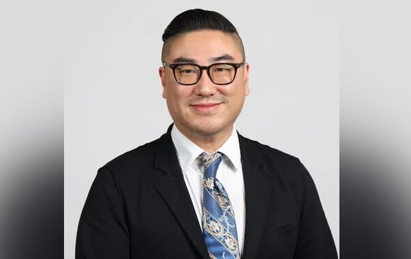 A headshot of a man with short black hair and eyeglasses. He wears a suit