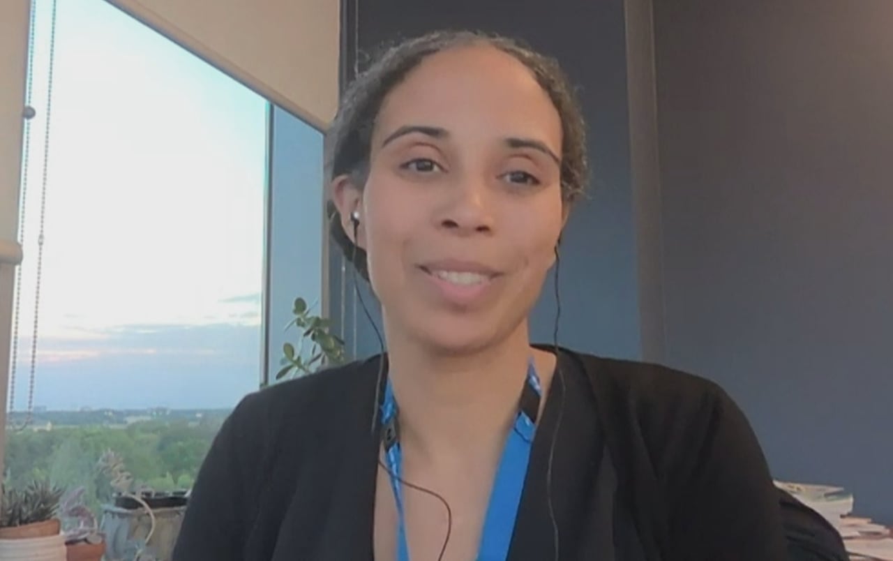 A woman looks into the camera. She sits at a table with a window behind her. She has curly hair pulled back in a bun.