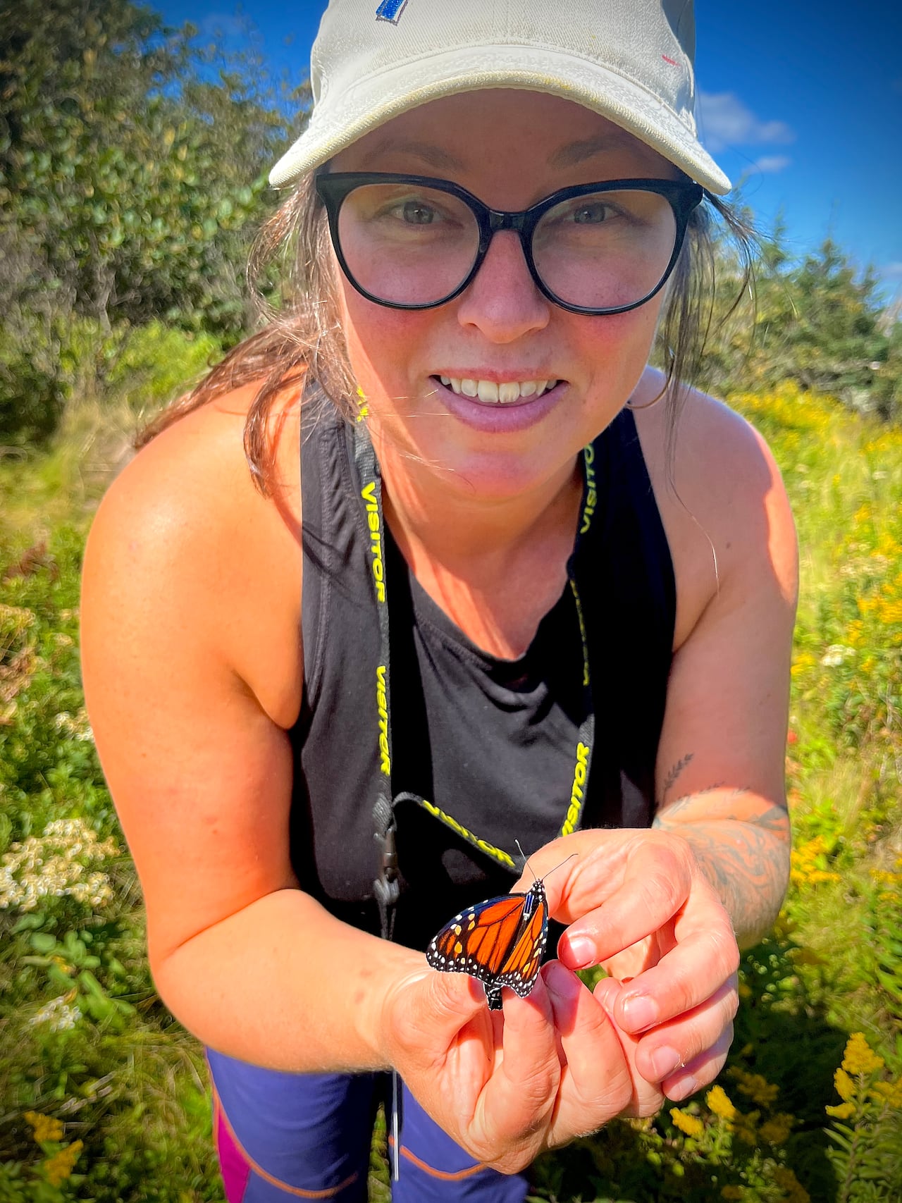 A smiling woman wearing glasses and a white cap holds a monarch butterfly with a small tag on its thorax close to the camera in a field of yellow wildflowers.