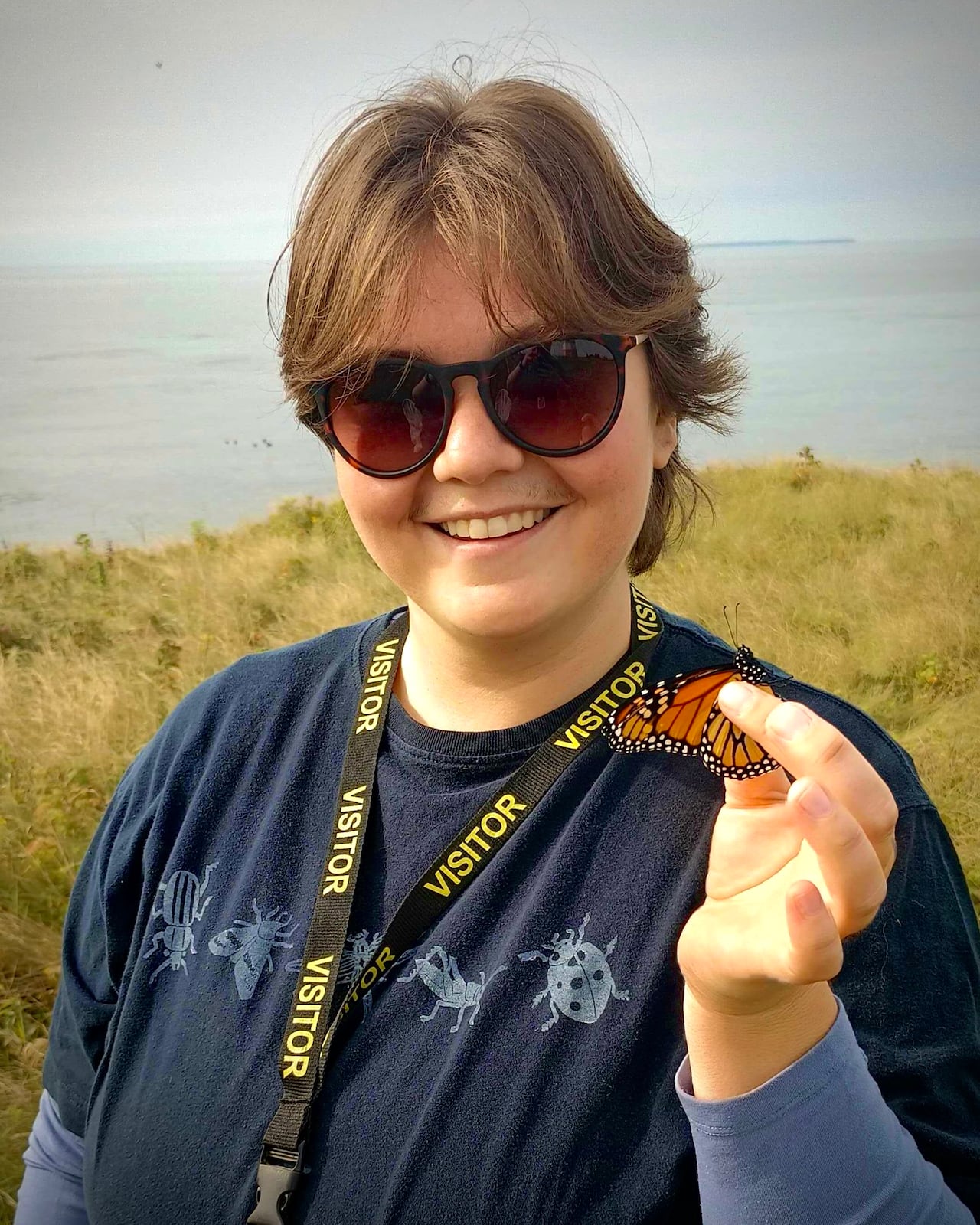 Woman wearing sunglasses and a visitor lanyard smiles while holding a monarch butterfly on her finger with the ocean and grassy dunes behind her.