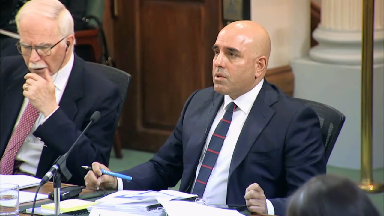 A man in a suit is seated at a long table. He has a stack of papers and microphone in front of him. He is holding a pen.