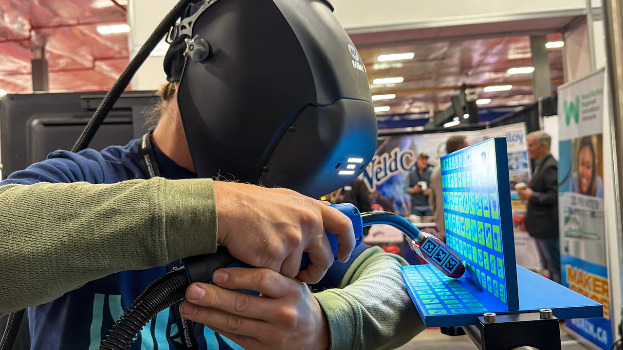 A man wears a welding helmet while demonstrating how to use a welding simulator at a trade show.