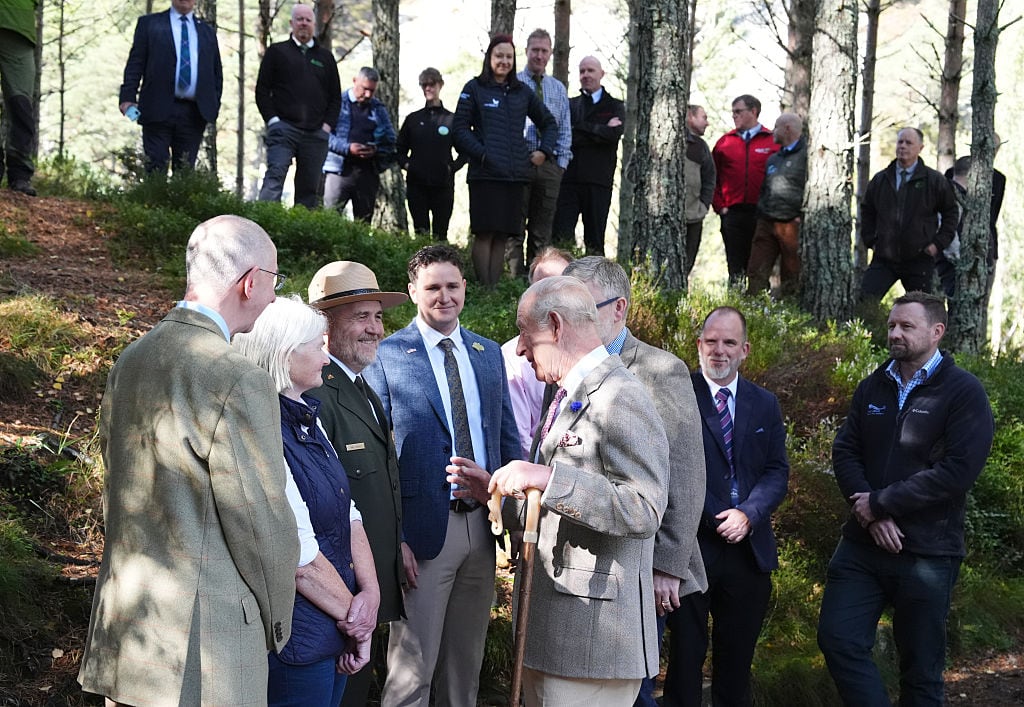 Several people watch as two people talk with one another along a path in a wooded area.