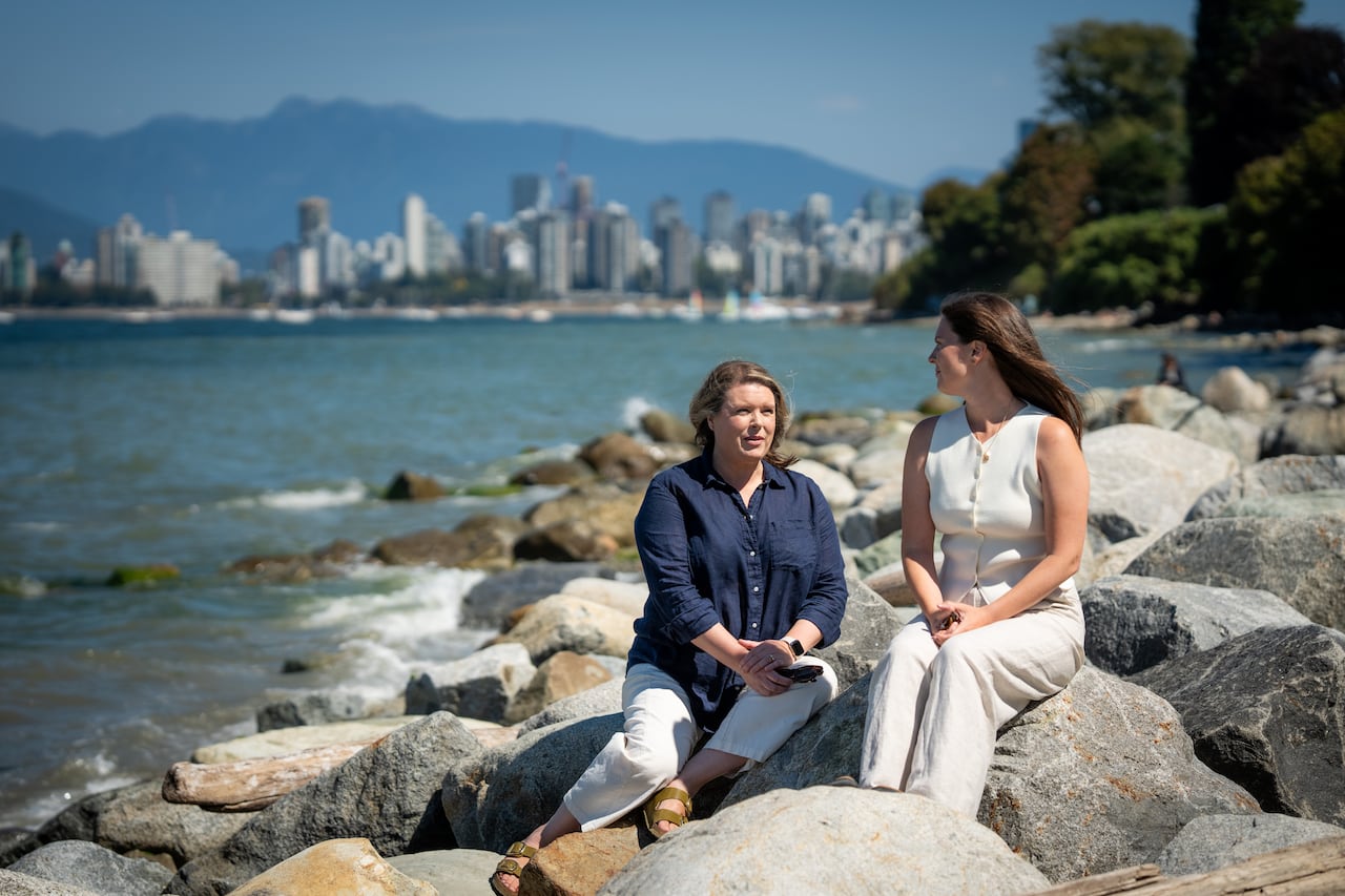 A woman in a blue shirt sits with a woman in a white blouse talking on rocks near the ocean with Vancouver in the background.
