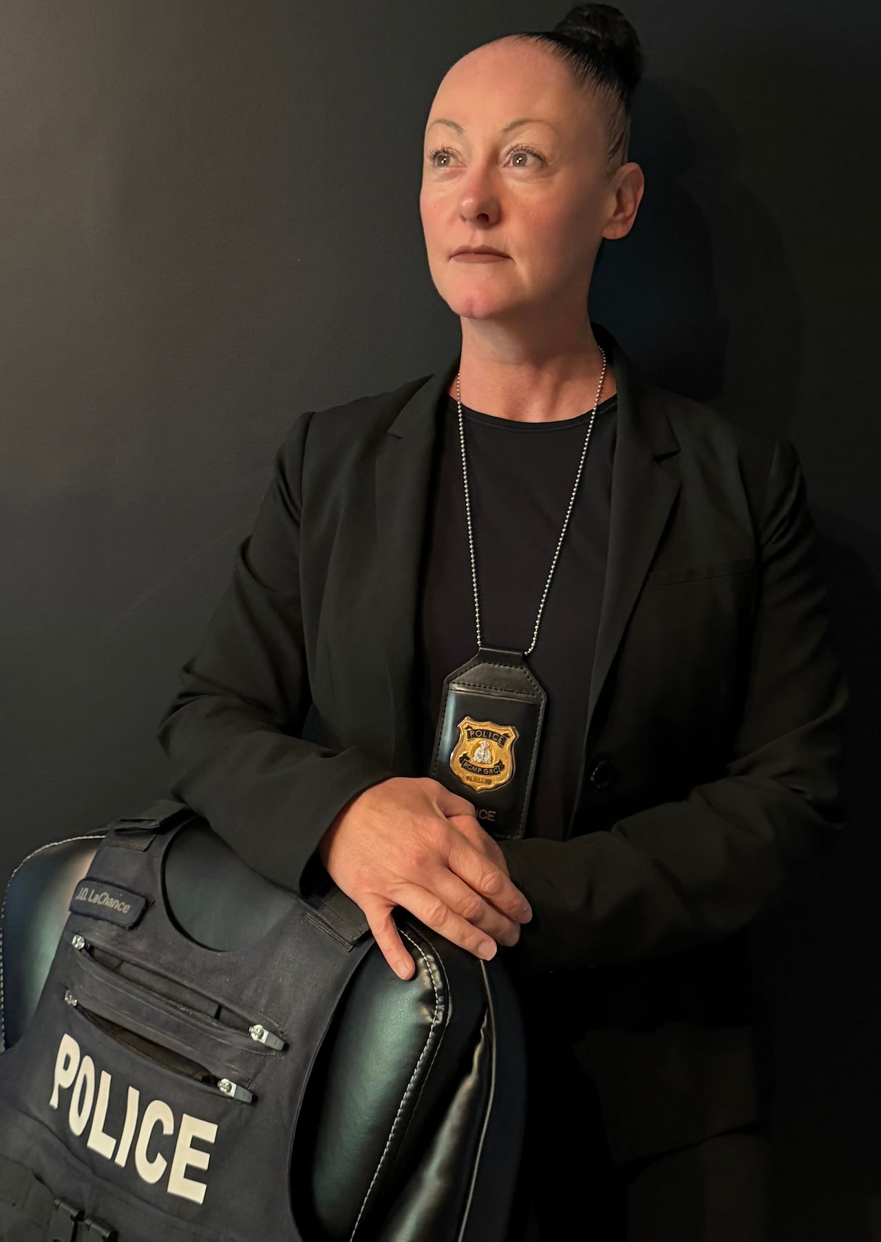 A woman with a dark shirt, police badge, and police vest stands beside a chair.