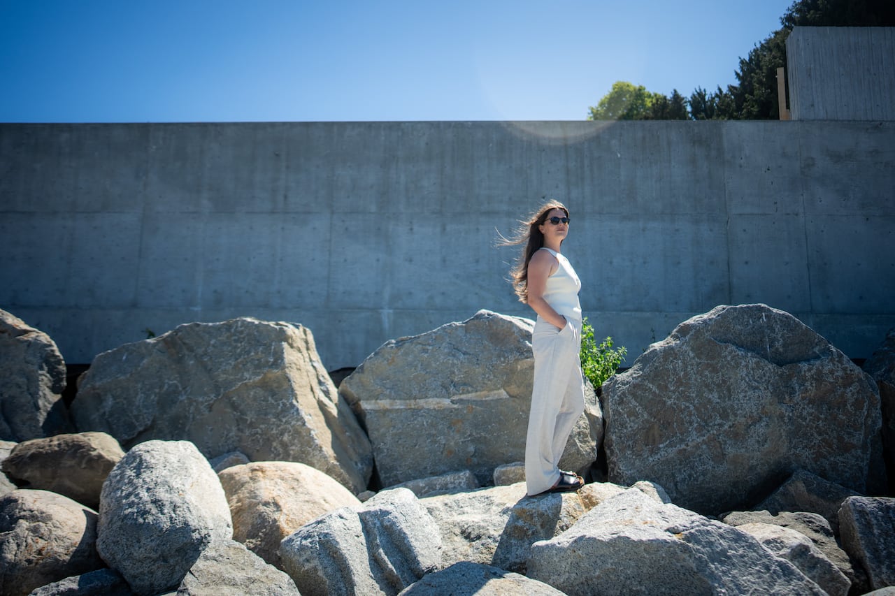 A woman wearing white walks in the breeze along some rocks at the beach.