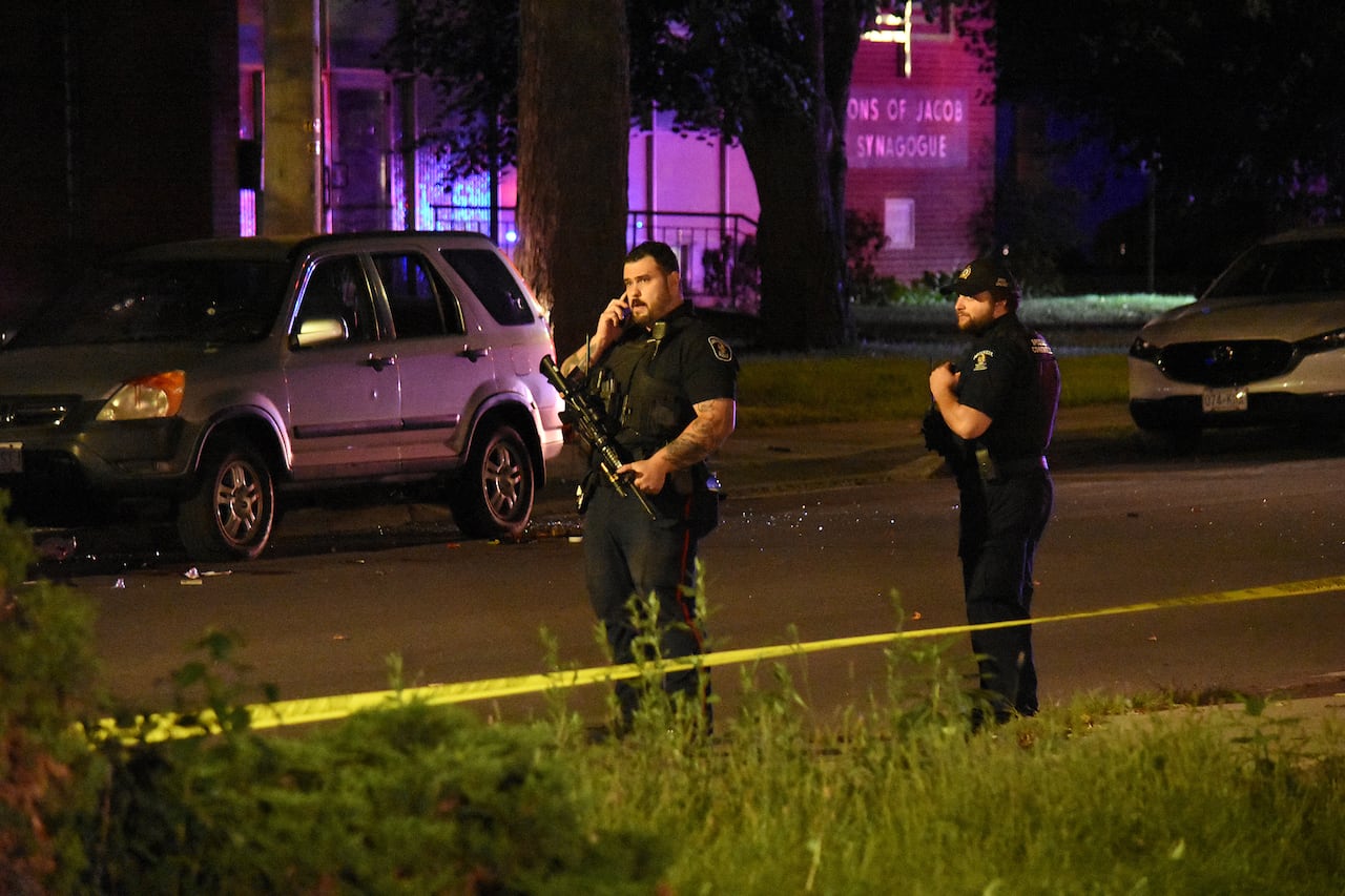 Two police officers stand behind police tape on the street at night outside a synagogue. One police officers speaks on a cellphone.