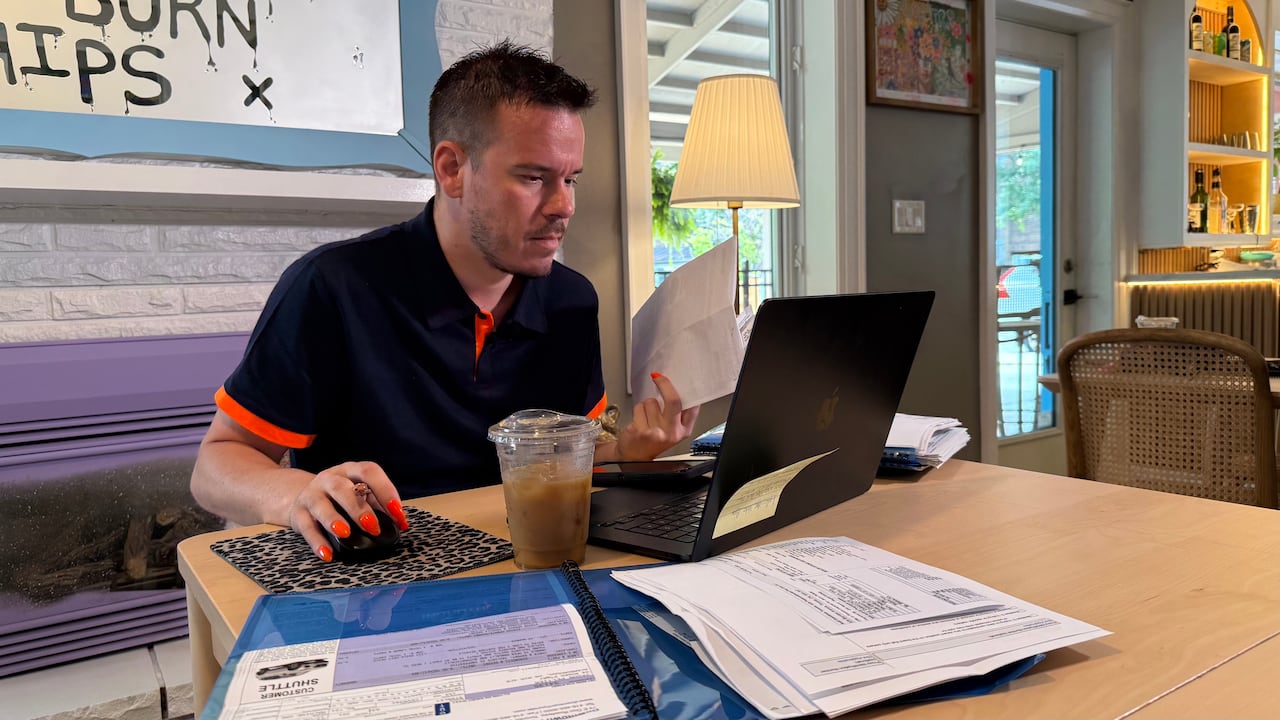 a man with dark hair sits in front of a laptop and sorts through paperwork