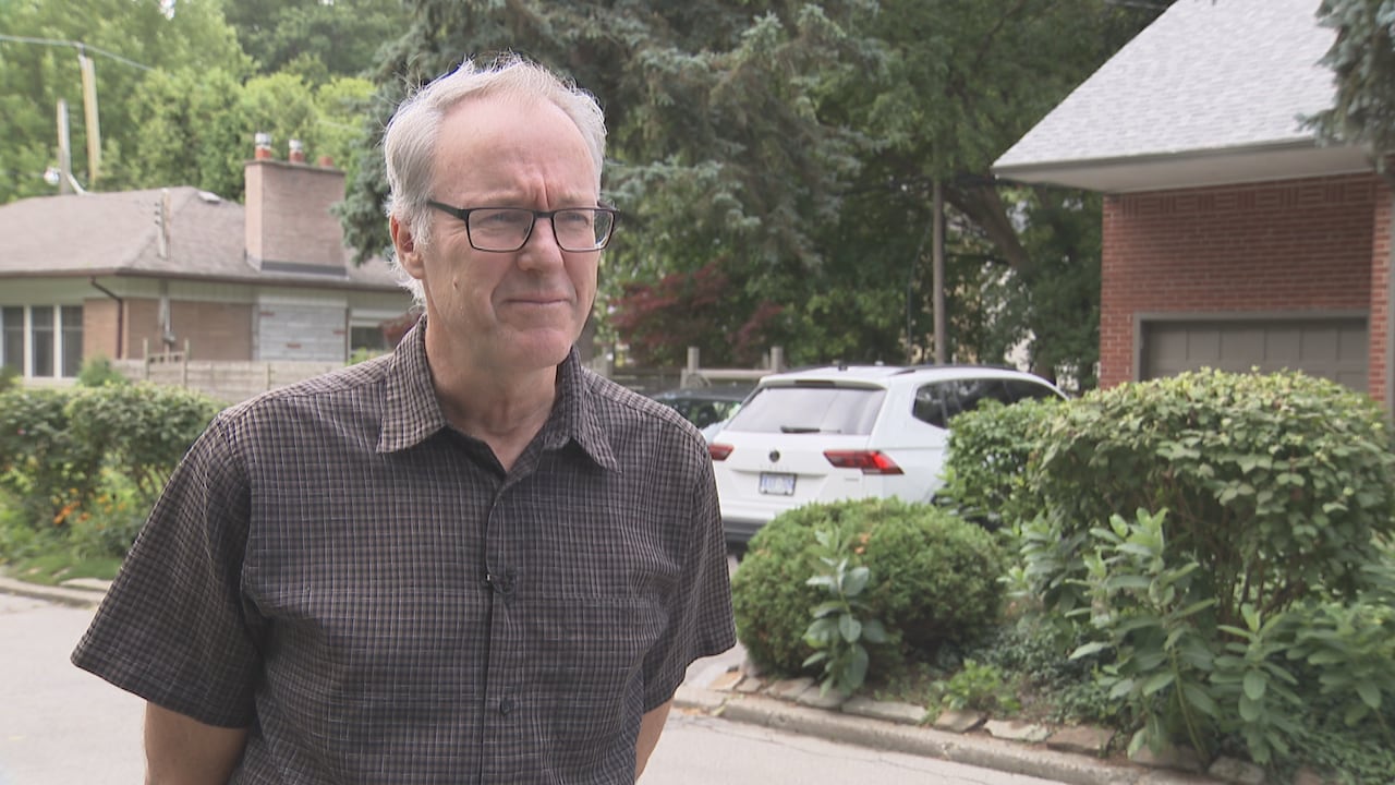 a man with grey hair and glasses and a plaid shirt stands on a residential street, speaking to someone off camera