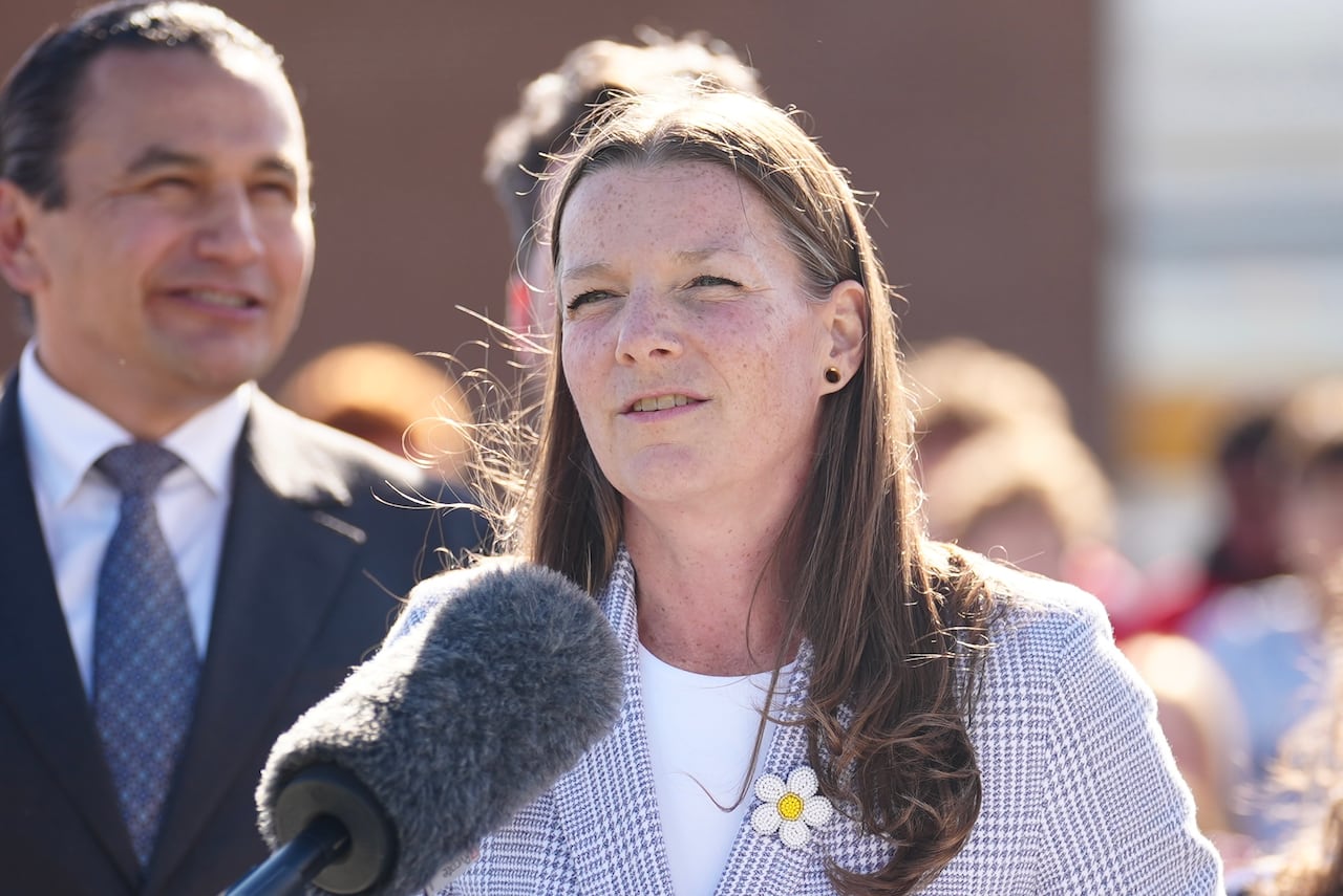 A woman with long hair squints in the sun as she speaks outdoors at a podium.