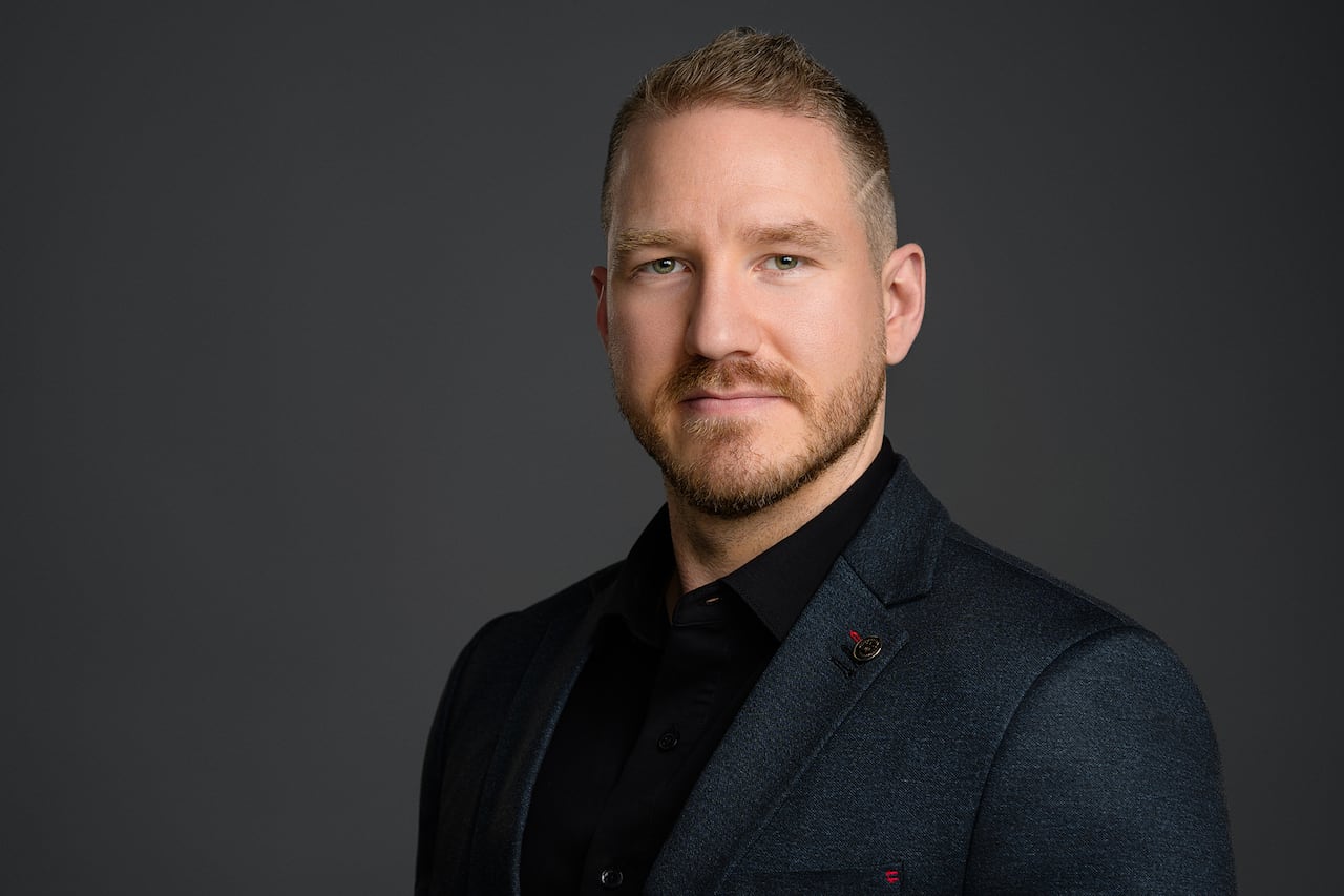 A man wearing a dark blazer and shirt poses for a headshot against a black backdrop.