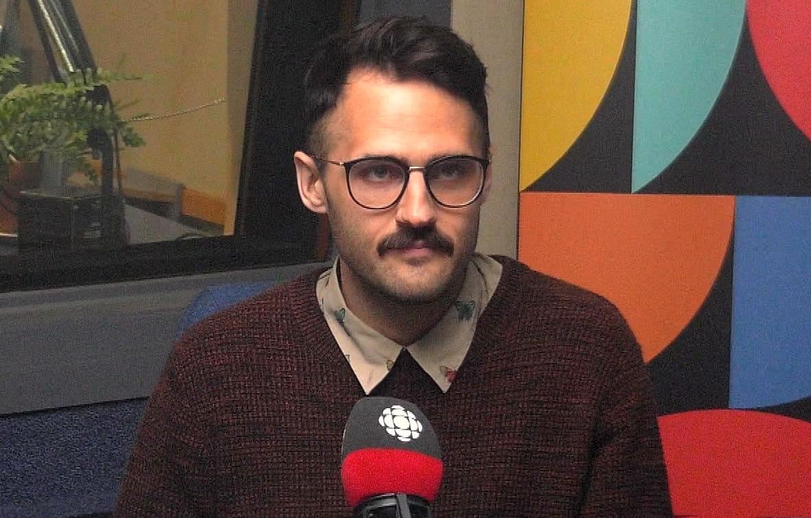 A man in glasses and a sweater sits in front of a microphone with the CBC logo on it.
