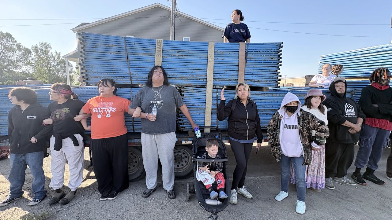A group of people link arms in front of a truck carrying stacks of blue fencing.