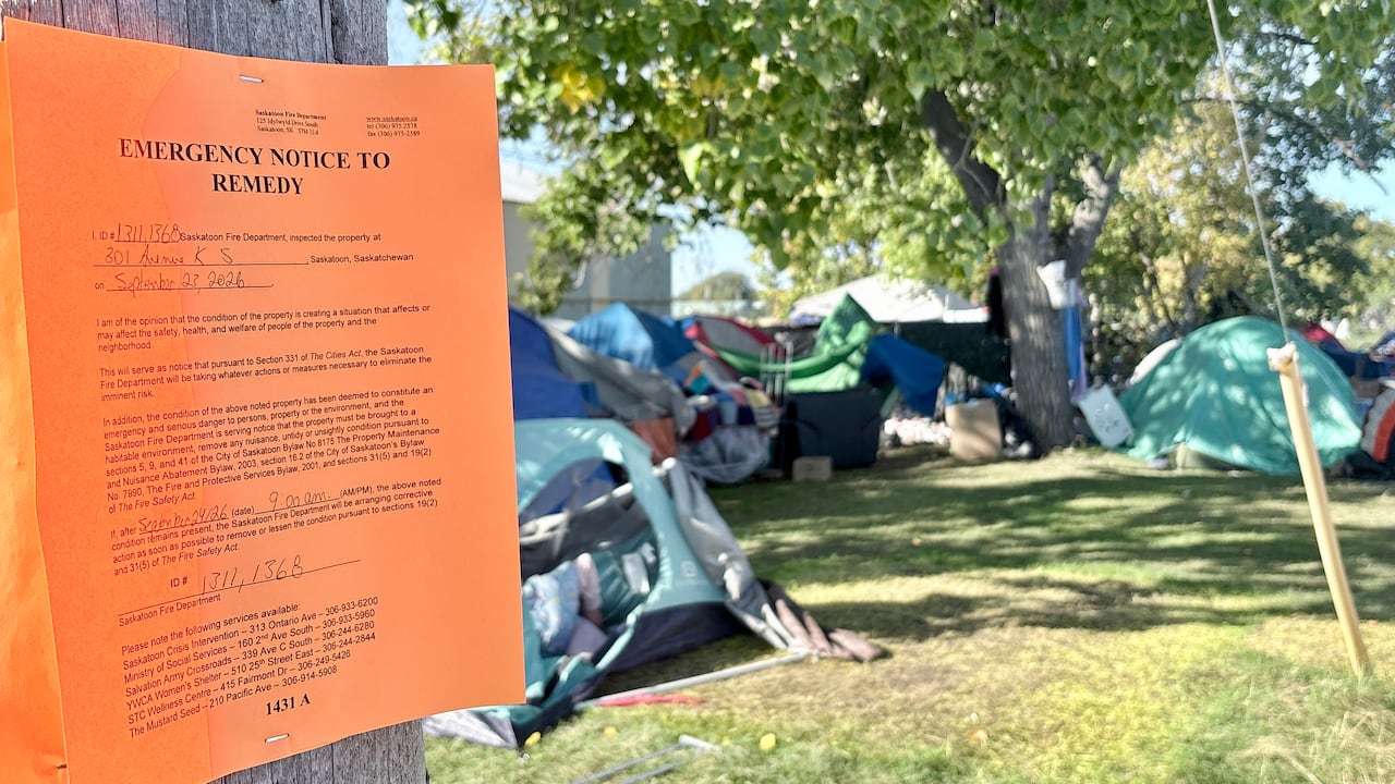 A piece of orange paper with the header "emergency notice to remedy" is affixed to a pole in front of a group of tents in a grassy area.