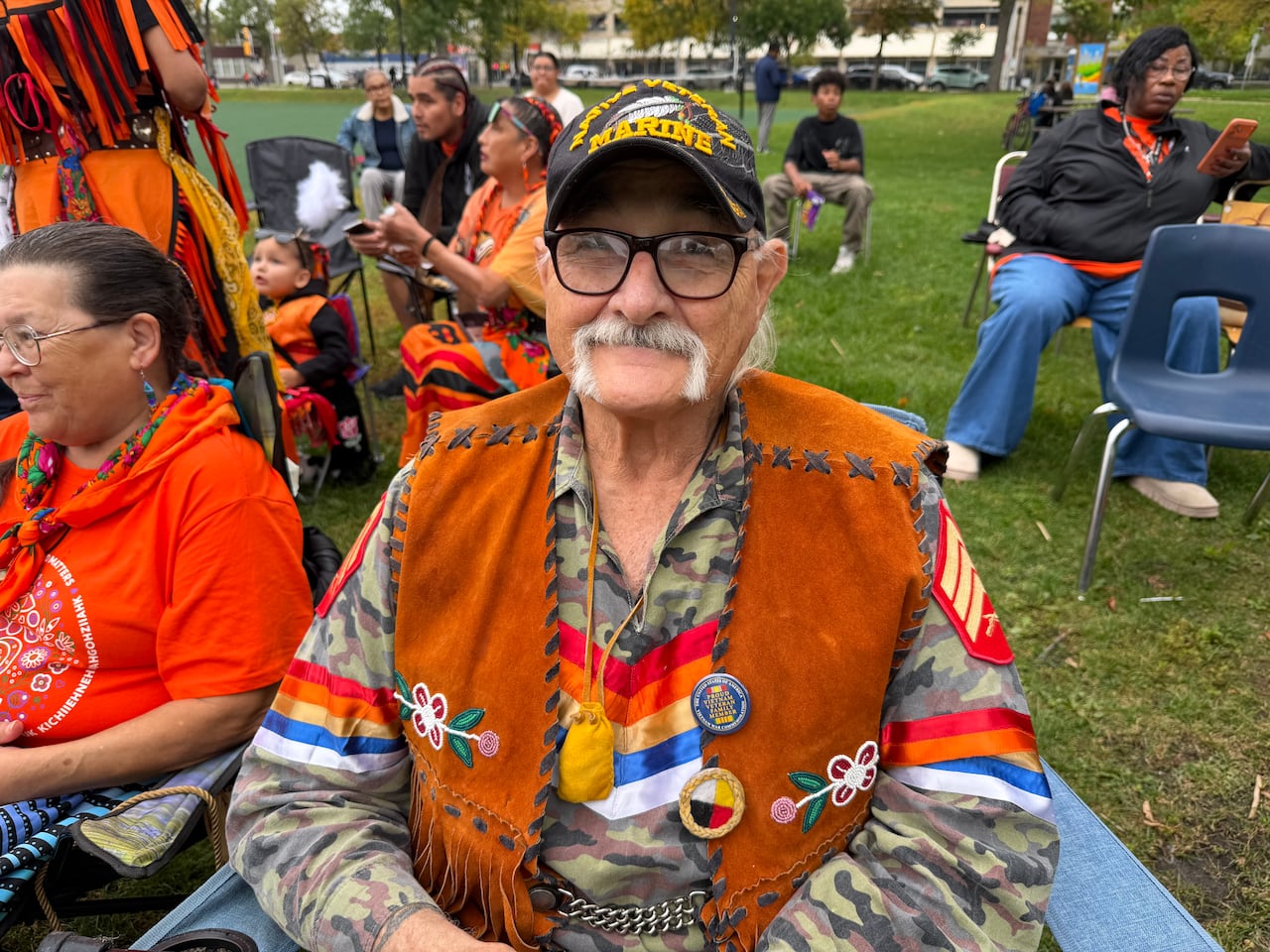 Man with a mustache wearing glasses and a baseball hat is sitting in a chair at the outdoor event.