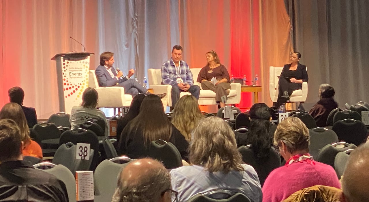 Two men and two women on a stage sitting down in front of a crowd of people sitting down in a convention centre room.