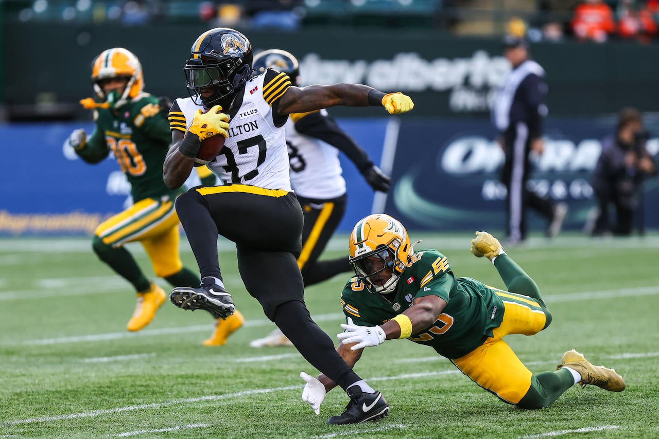 A men's football player, wearing green-and-yellow, dives toward an opponent, wearing white-black-and-yellow, who is running with the ball.