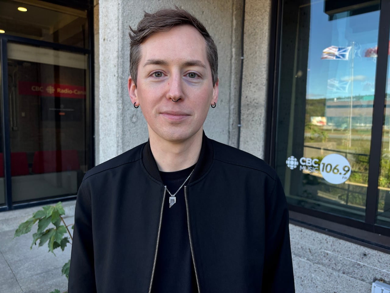A person with short hair and black jacket stands in front of a window.