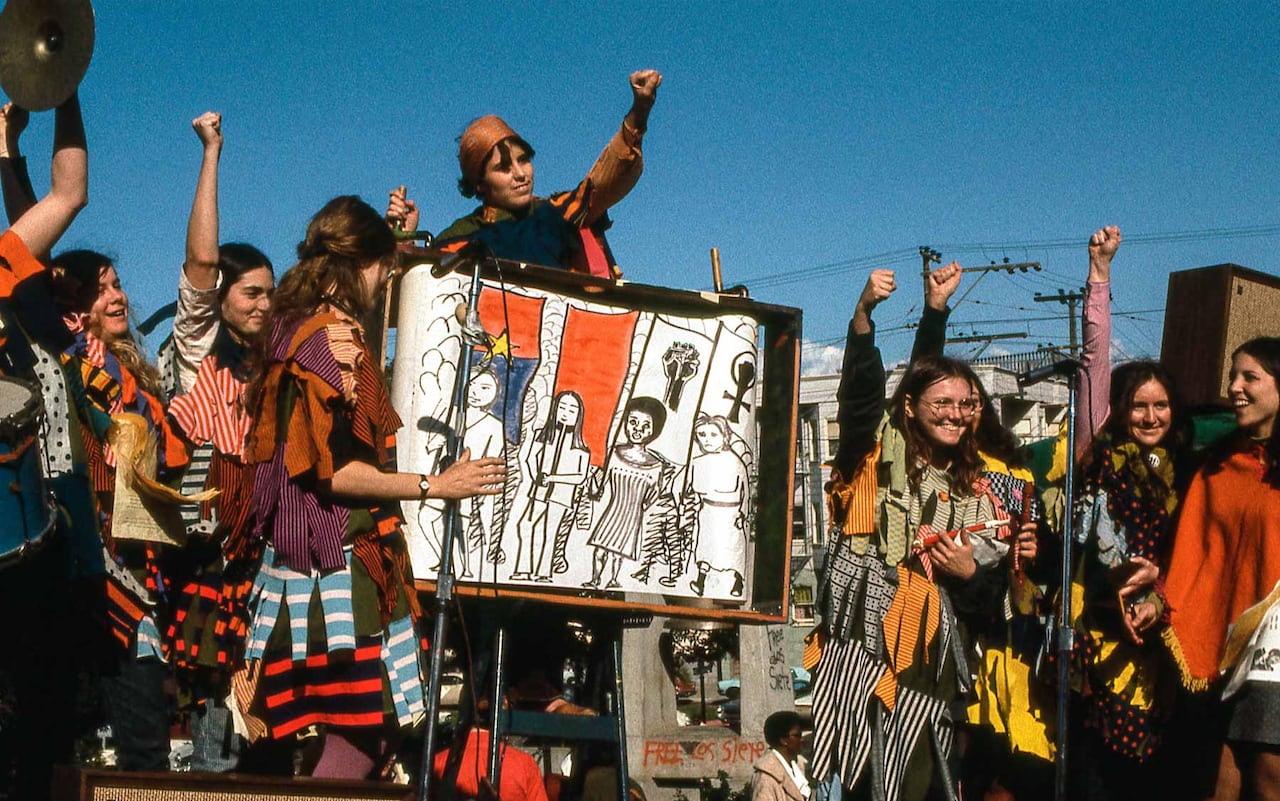 A group of people in colourful dress stand with their arms raised, with the person in the centre holding artwork depicting four people drawn out.