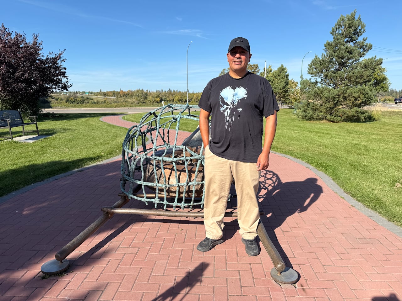 A tall First Nations man in a ballcap, black t-shirt and tan pants stands by a bronze sculpture in a park in the summer.