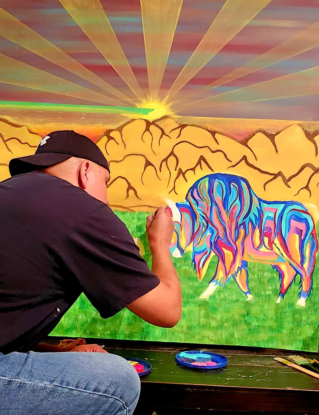 A First Nations man in a black t-shirt and ballcap sits painting a canvas with a sunset and two buffalos.