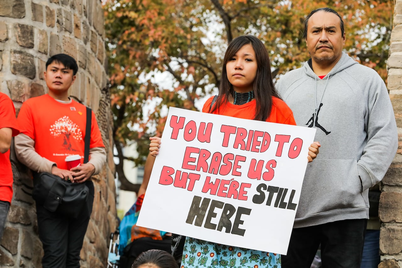 A young woman wearing an orange shirt and a skirt holds a sign reading, 'You tried to erase us, but we're still here.' Others in orange shirts stand behind her.