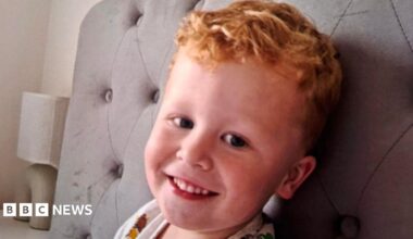 Three-year-old Noah Woods, with light hair and wearing a brightly coloured top, smiles at the camera.