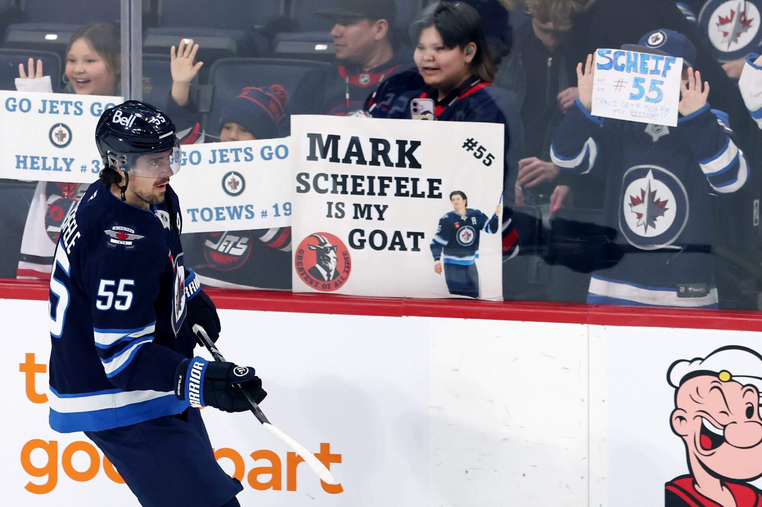 Mark Scheifele, with his uniform number of 55 on his right shoulder, skates past fans, some of whom are holding signs, before a game.
