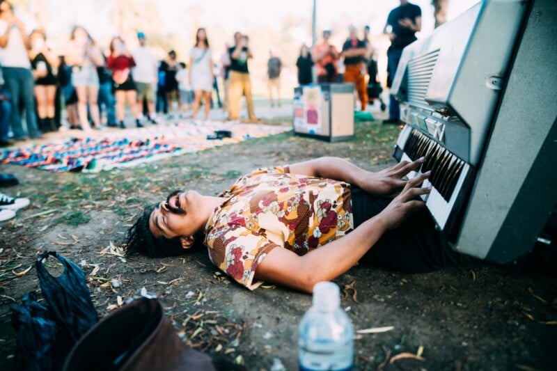 A man with a mustache lies on the ground, playing a keyboard positioned vertically above him, while a crowd watches in the background.