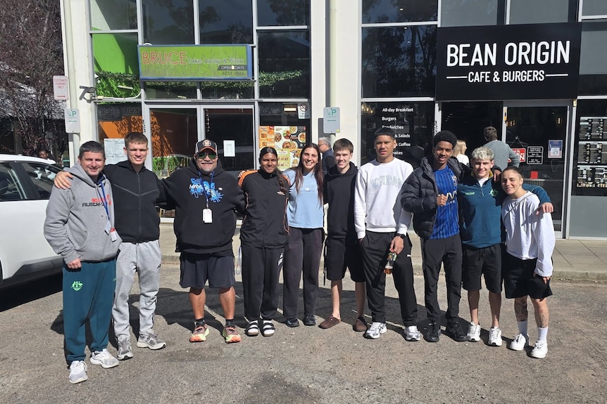 A group of 10 people, 1 adult and 9 teenagers, line up together smiling for the camera outside a cafe.