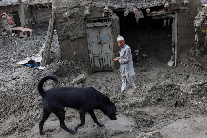man walks in front of a mud-caked house with a black dog in the foreground