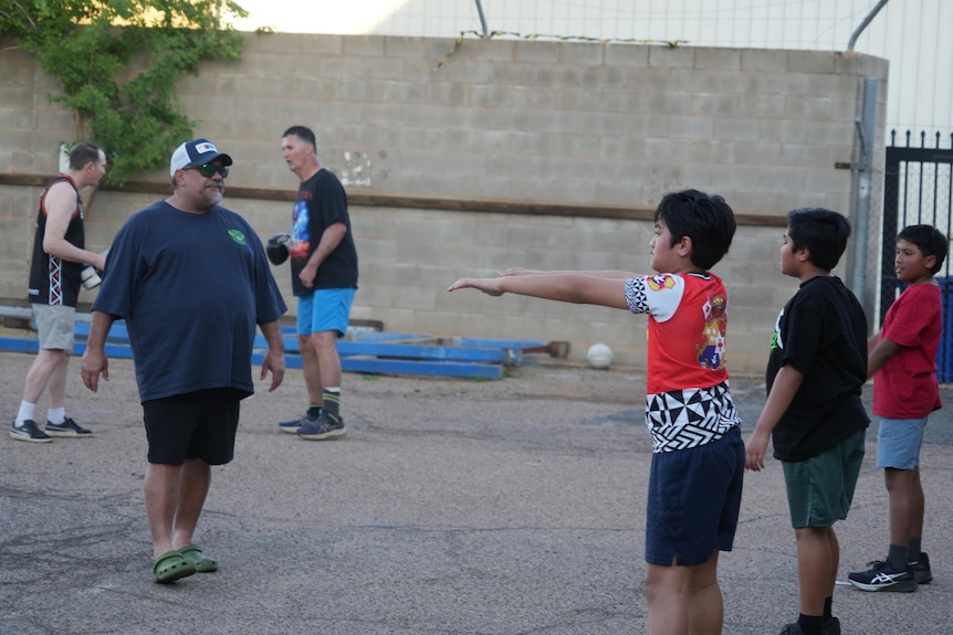 A man in a baseball cap faces a line of three children performing exercises.