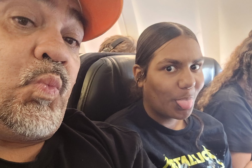 Sat on an aeroplane, a bearded man pulling a kissing face sits alongside a 12-year-old girl poking her tongue out.