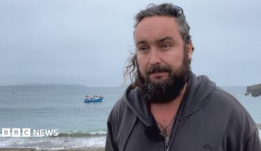 Deputy Lee van Katwyk has long hair tied back and a full beard. He is standing on a beach wearing a dark zip-up hoodie with a microphone clipped to the front. Calm sea water and small waves are visible behind him, with a blue fishing boat anchored offshore. A rocky headland extends into the water on the right under an overcast, grey sky.