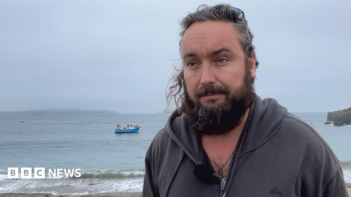 Deputy Lee van Katwyk has long hair tied back and a full beard. He is standing on a beach wearing a dark zip-up hoodie with a microphone clipped to the front. Calm sea water and small waves are visible behind him, with a blue fishing boat anchored offshore. A rocky headland extends into the water on the right under an overcast, grey sky.
