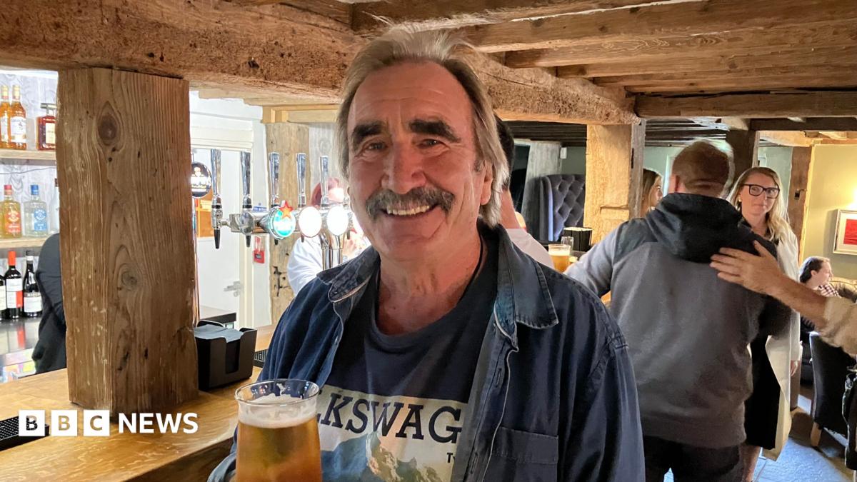 A man with a moustache. He is holding a crisp pint.