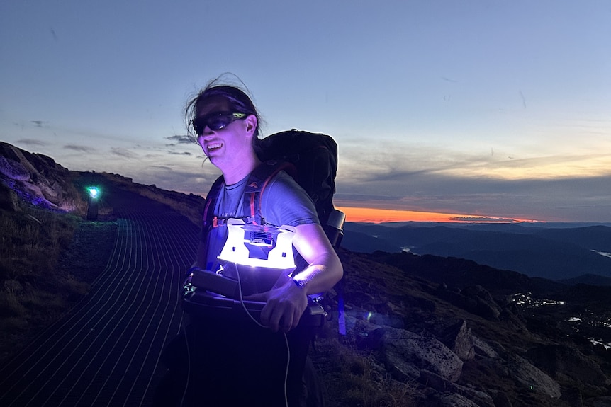 A man in a t-shirt carrying a large backpack and an illuminated lantern, standing on a mountain path