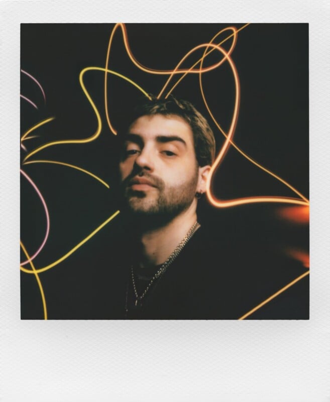 A young man with short brown hair and a beard looks directly at the camera against a black background featuring abstract, glowing yellow light trails.