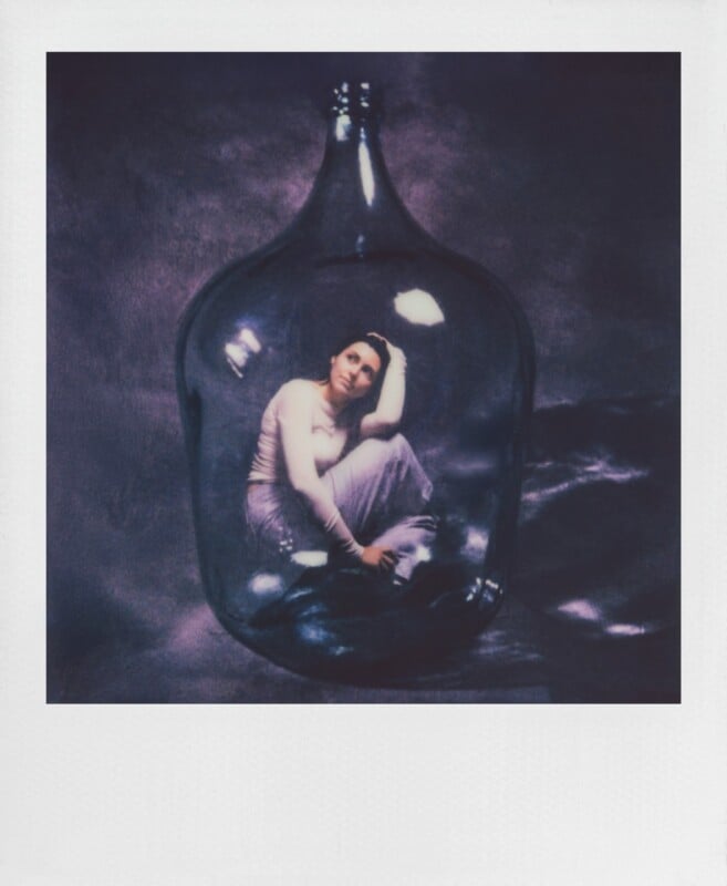 A person sits curled up inside a large, clear glass bottle against a dark, textured purple background.