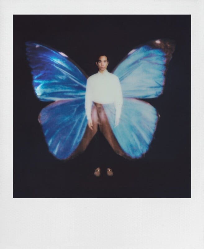A person stands centered against a black background, wearing a white shirt and dark pants, with large, glowing blue butterfly wings superimposed behind them.