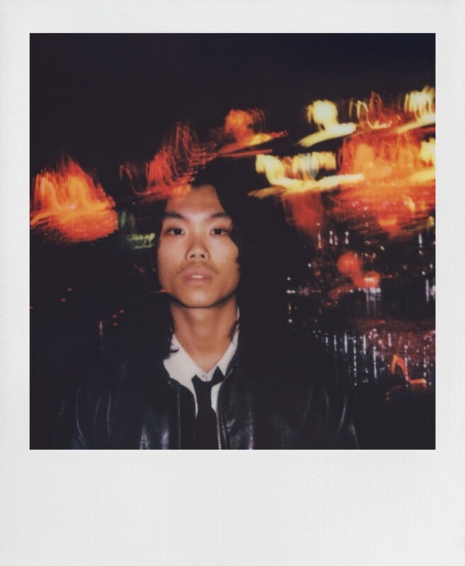 A young person with long dark hair, wearing a black leather jacket and a white shirt with a black tie, stands against a dark background with blurred, glowing orange and yellow light streaks.