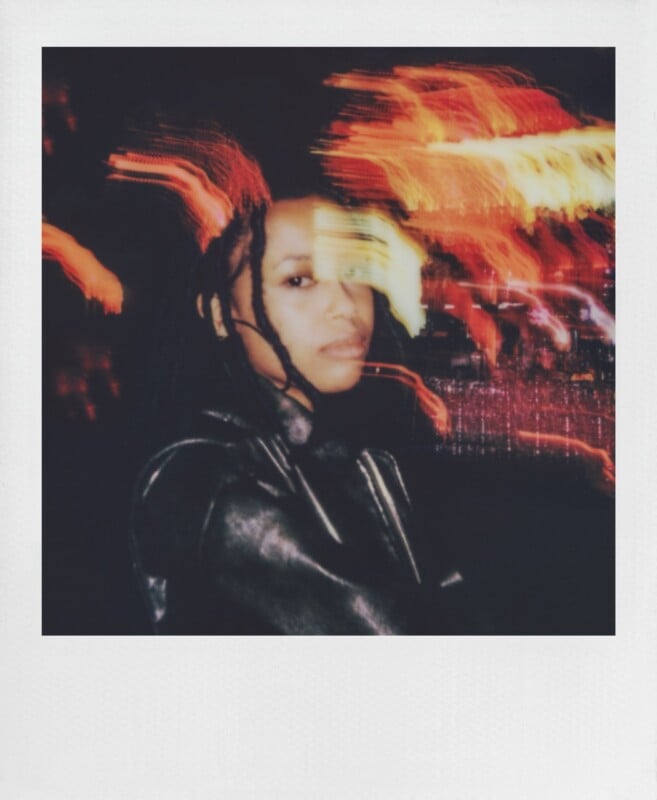 A person with dark hair and a leather jacket looks toward the camera against a dark background with blurred, streaking lights.