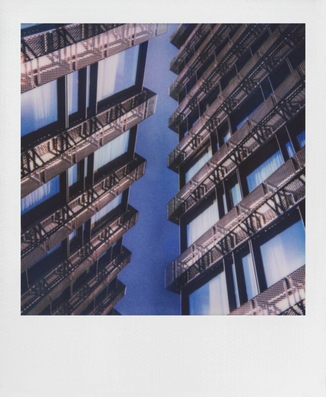 Two modern buildings with dark, perforated metal facades and large windows face each other against a clear blue sky.