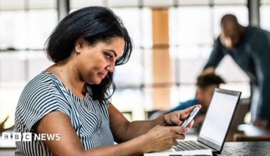Woman looking at a phone sits at a desk with a laptop in front of her and a man and boy in the background.