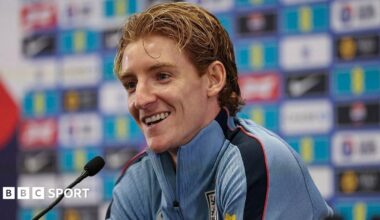 A smiling Anthony Gordon, in a blue zip-up top, sits by a microphone at an England news conference