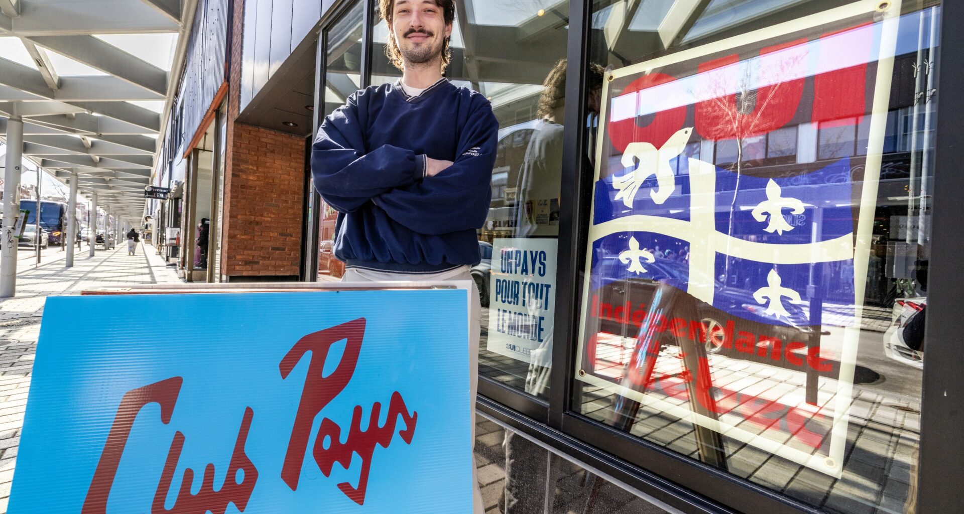 Man stands outside cafe window decorated, which is decorated with a Quebec flag, with a sovereignty sign on the sidewalk in front of him.