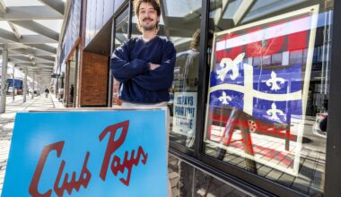 Man stands outside cafe window decorated, which is decorated with a Quebec flag, with a sovereignty sign on the sidewalk in front of him.