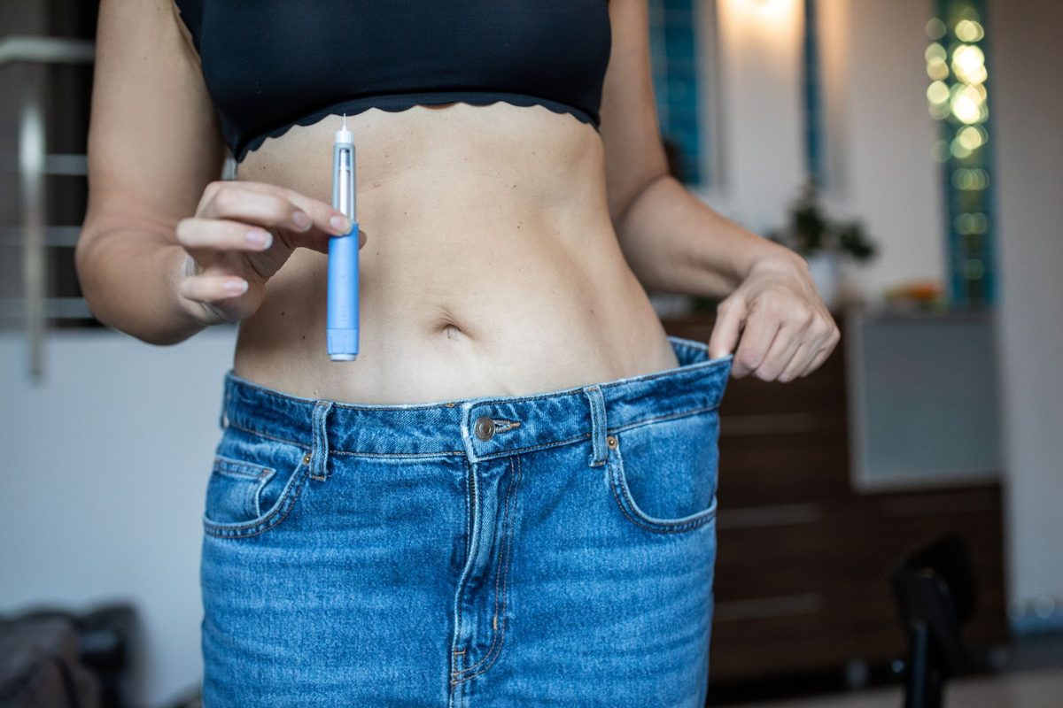 A person in jeans that are too big holds the waistband in one hand and an injector pen in the other